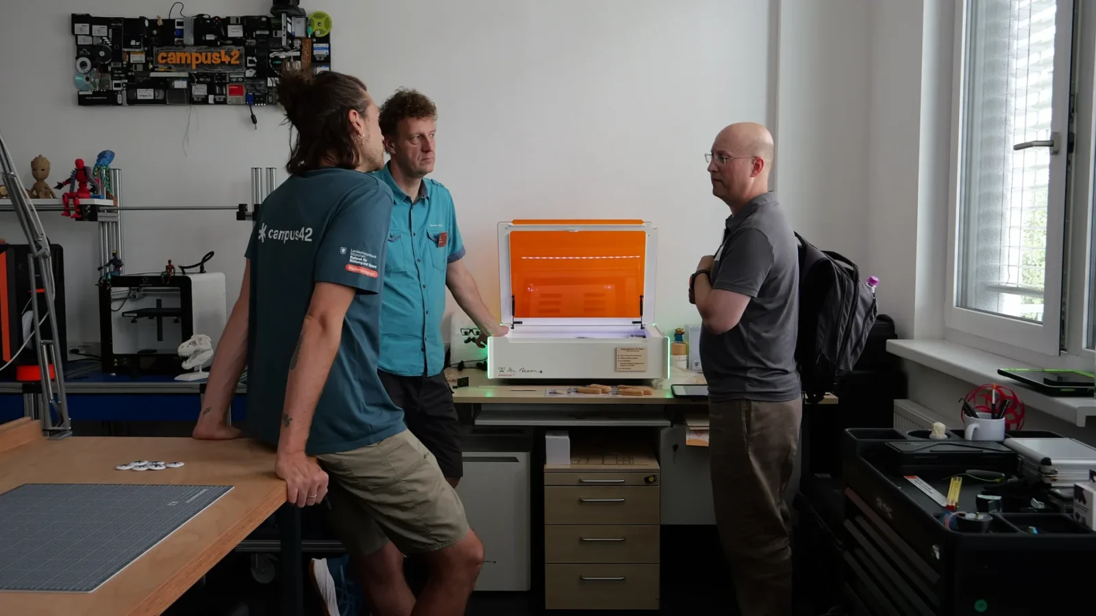 Drei Personen im Makerspace vor einem geöffneten Lasercutter, im Hintergrund 3D-Drucker und campus42-Logo