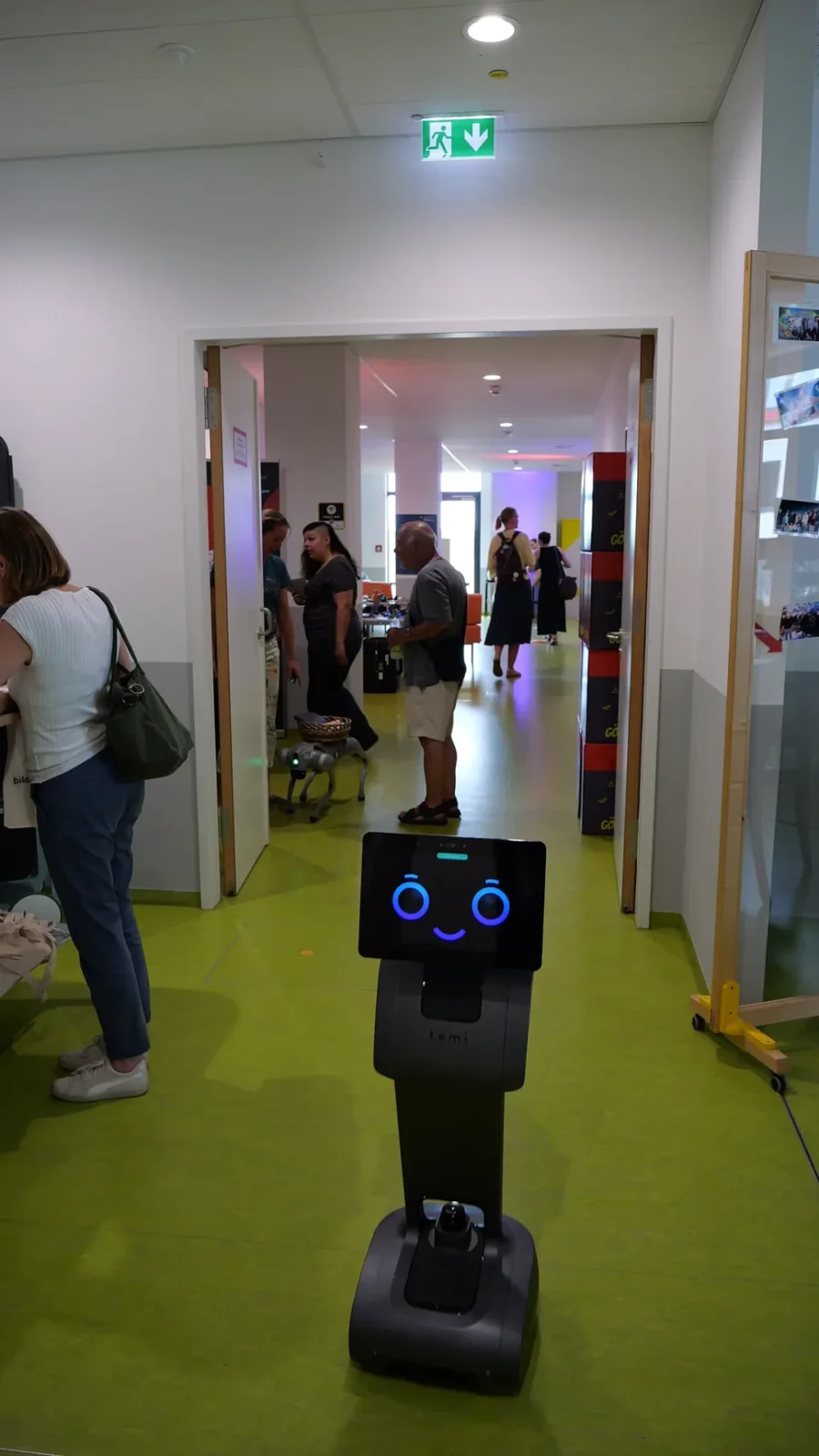 Servierroboter mit Display-Gesicht im Flur, dahinter ein Roboterhund und Besuchende