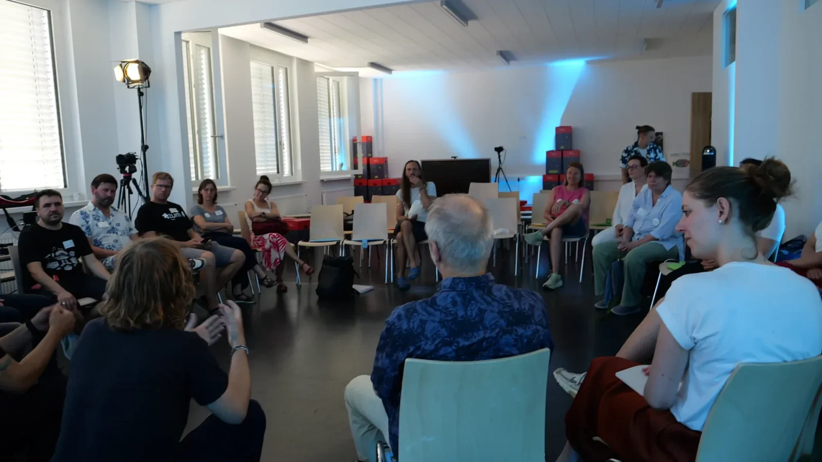 Barcamp-Session im Stuhlkreis, Teilnehmende diskutieren neben Filmscheinwerfer und Kamerastativ