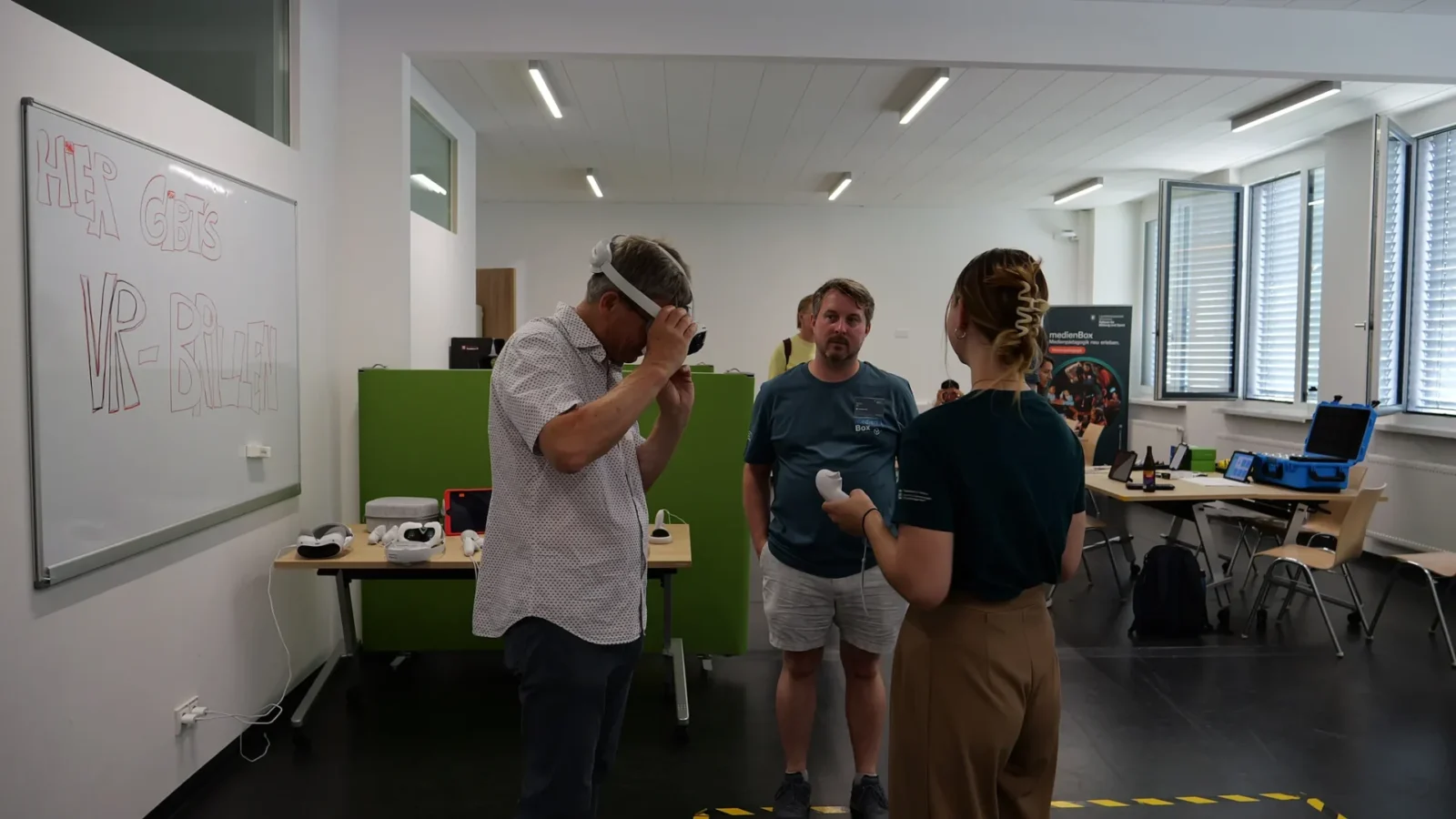 Besucher setzt eine VR-Brille auf, daneben das Whiteboard „Hier gibt's VR-Brillen" und ein medienBox-Rollup